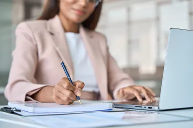 Close up of woman in pink blazer writing with computer open